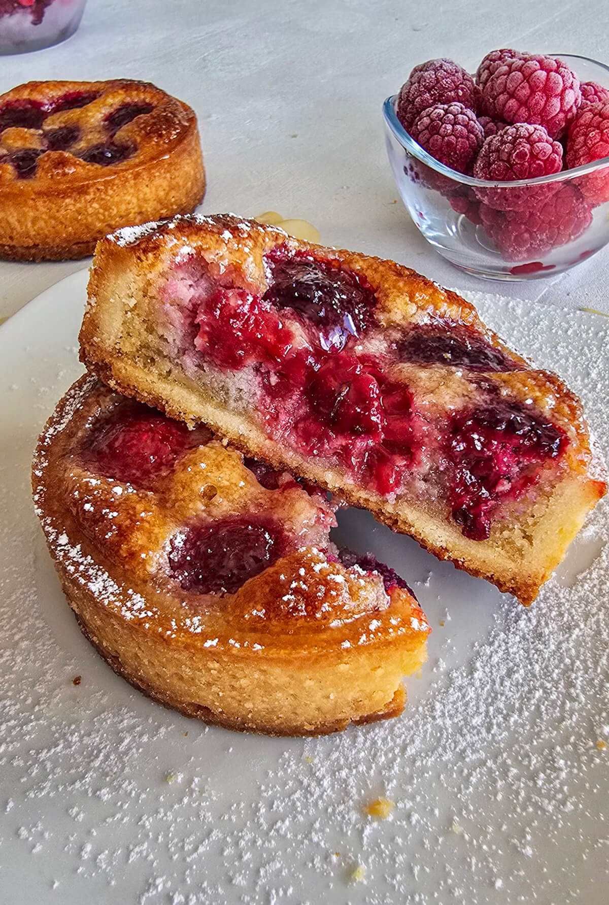 Une tartelette framboise coupé en deux, sur une assiette blanche, framboise en deuxième plan.