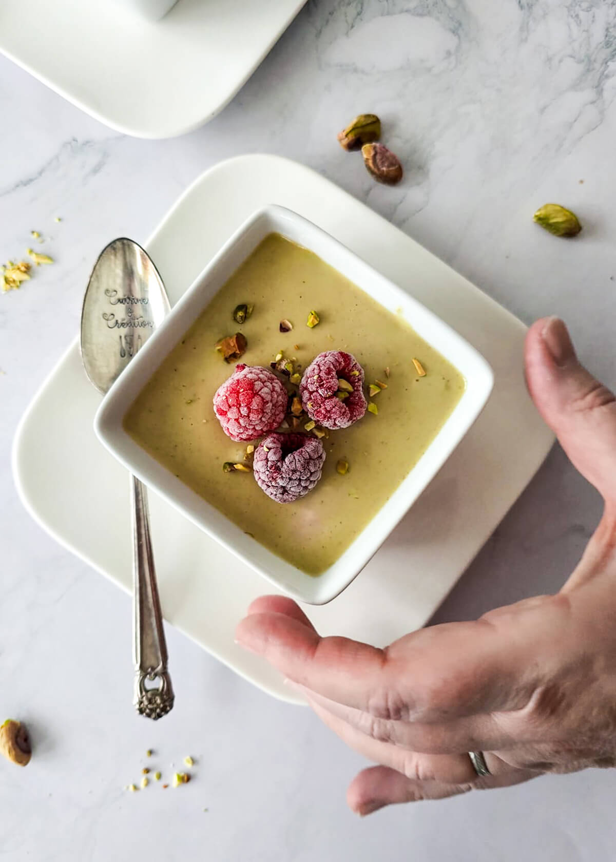 Une main tenant une verrine remplie de crème dessert pistache avec des framboises.