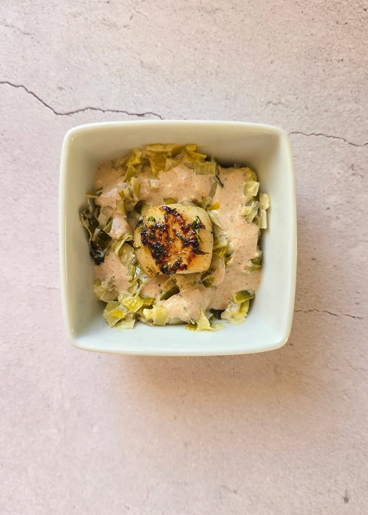 Une cassolette de saint Jacques et sa fondue de poireaux dans un ramequin blanc.
