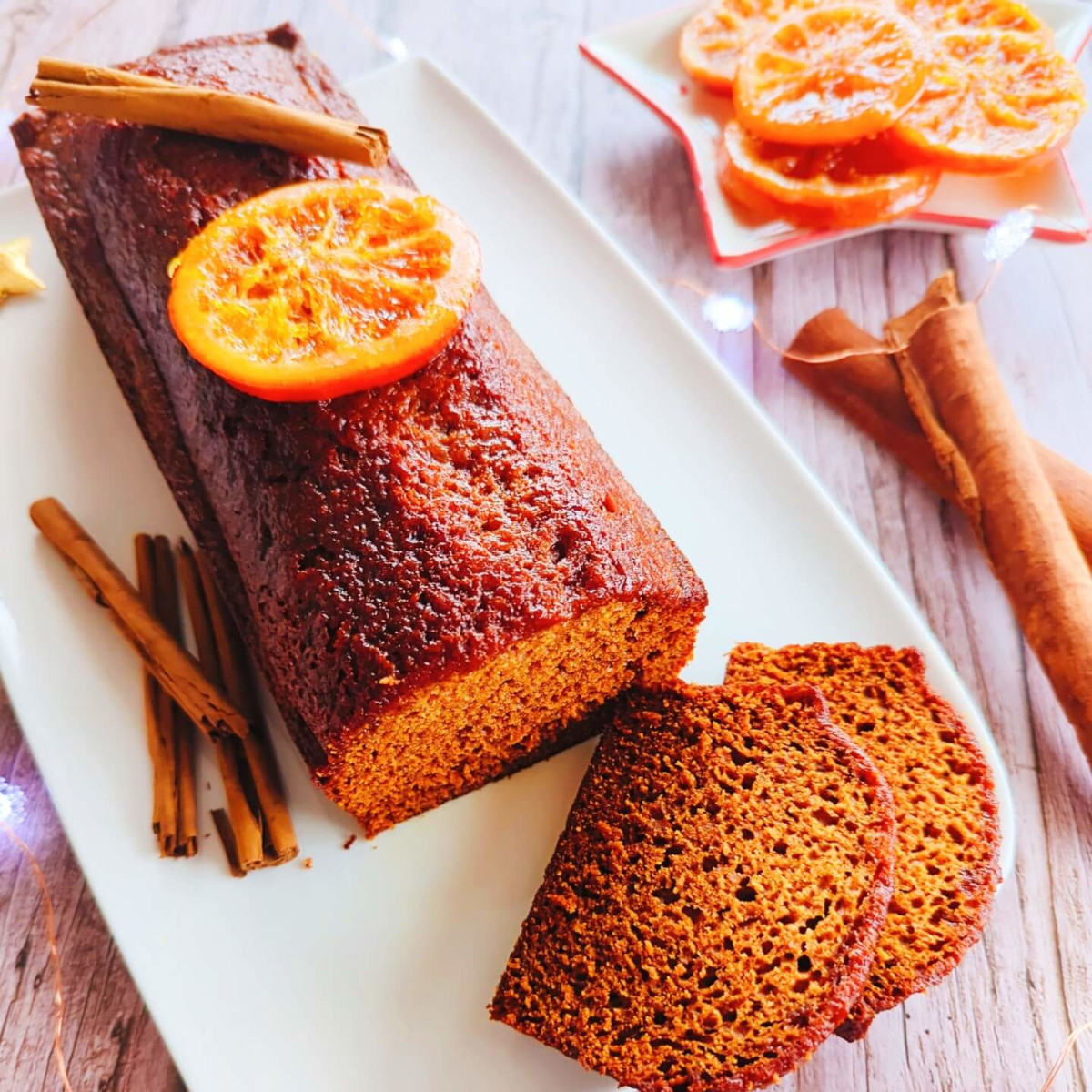 slices of traditional gingerbread on a white serving plate with cinnamon and candied orange slices, set against a wooden background with fairy lights.