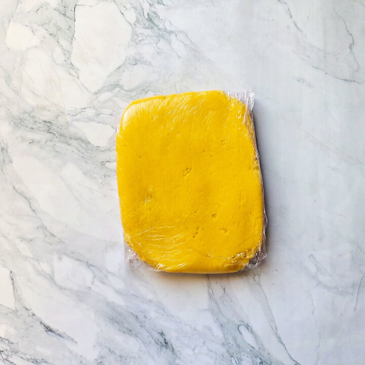 Pâte à palets bretons façonnée en carré et enveloppée dans du film alimentaire, posée sur un plan de travail en marbre pour le temps de repos au frais.