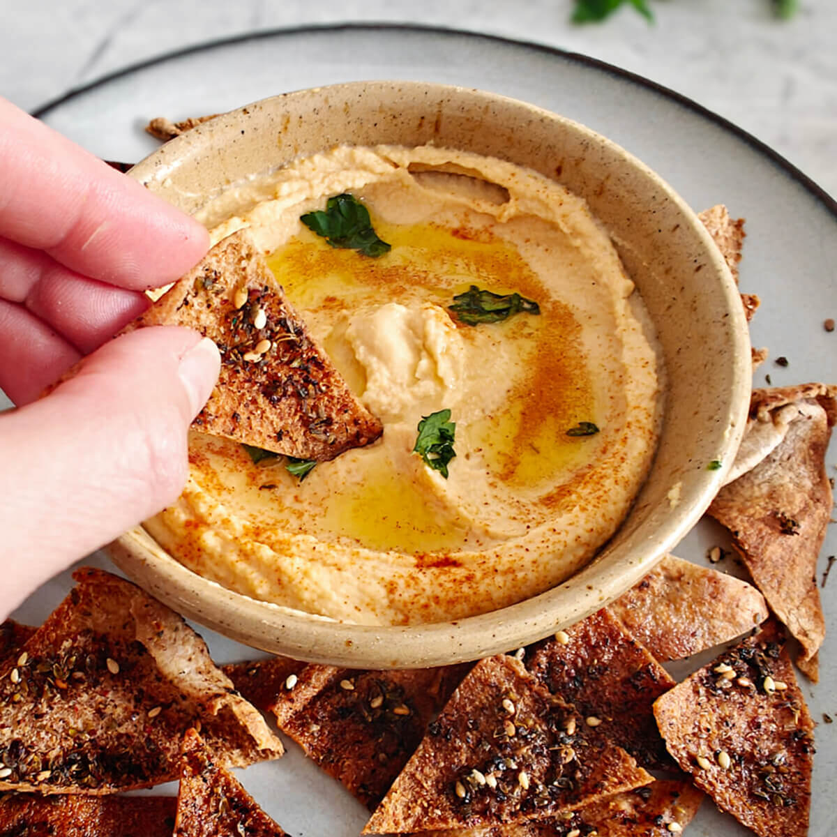 Un houmous bien crémeux dans un bol, avec une main tenant une chips de pita.
