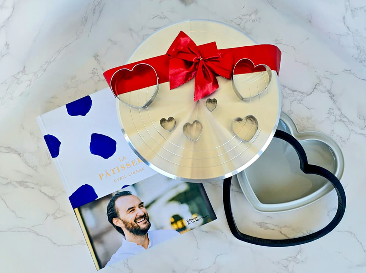 Sélection d'idées cadeaux pour la fête des mères : le livre de cuisine "La Pâtisserie" de Cyril Lignac, accompagné d'un moule à gâteau en forme de cœur et d'emporte-pièces assortis sur un plan de travail en marbre.