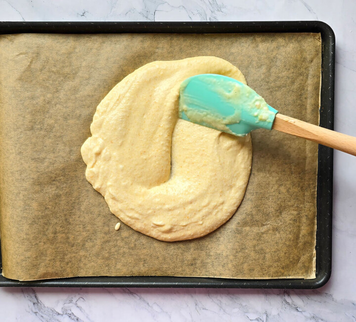 Pâte à biscuit Joconde étalée à l'aide d'une spatule souple sur une plaque de cuisson recouverte de papier sulfurisé.