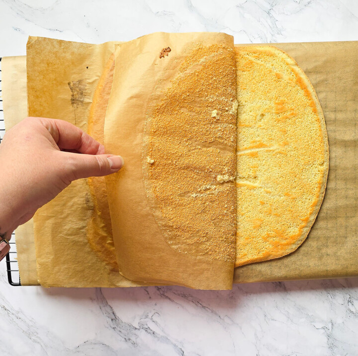 Une main décollant délicatement une feuille de papier cuisson d'un biscuit Joconde moelleux et doré posé sur une grille de refroidissement.
