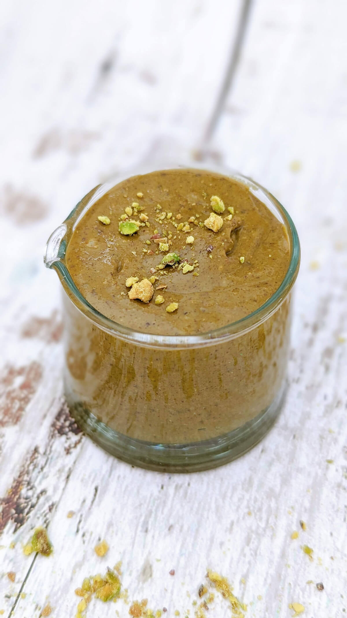 Du praliné pistache fait maison dans un petit contenant en verre, éclats de pistache concassées sur le dessus, sur un fond en bois clair.