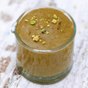 Du praliné pistache fait maison dans un petit contenant en verre, éclats de pistache concassées sur le dessus, sur un fond en bois clair.