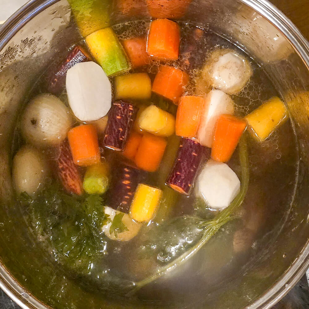 Ingrédients du pot-au-feu ajoutés au bouillon, incluant des morceaux de carottes orange et violettes, des navets et du céleri.