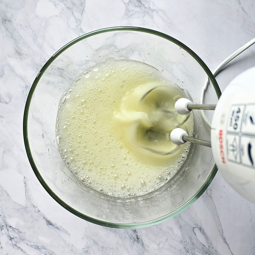Gros plan sur un batteur électrique en train de fouetter des blancs d'œufs dans un saladier en verre sur un plan de travail en marbre blanc.