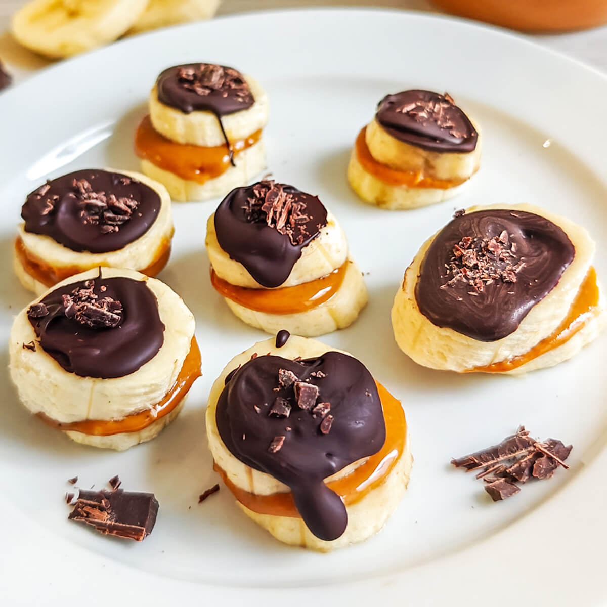 Bouchées de rondelles de bananes garnies de beurre de cacahuète crémeux et nappées de chocolat noir fondu, parsemées de copeaux de cacao sur une assiette blanche.