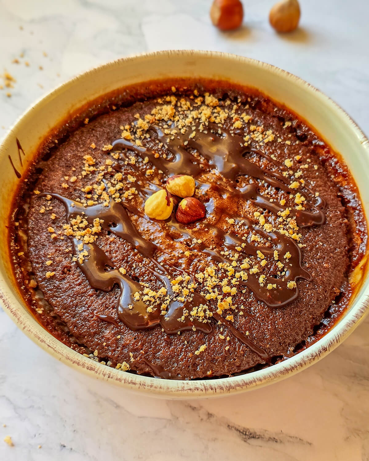 Alt text : Bowl cake au chocolat et noisettes parsemé d'éclats de noisettes et d'un coulis de chocolat fondant, servi dans un bol beige sur un plan de travail en marbre.