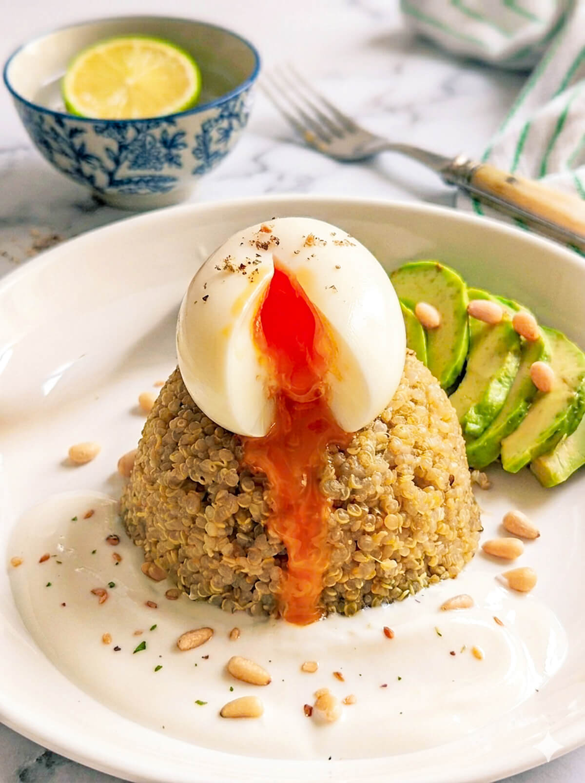 Gros plan d'un bowl sain composé de quinoa doré surmonté d'un œuf mollet au jaune coulant orange vif. À côté, des tranches d'avocat frais parsemées de pignons de pin et d'herbes sur une sauce crémeuse blanche. Présentation élégante sur une assiette blanche posée sur un plan de travail en marbre blanc, avec un bol de citron vert et un torchon à rayures en arrière-plan.