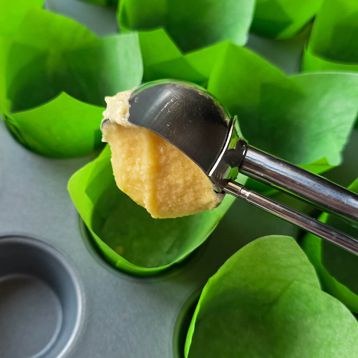 Main déposant de la pâte à cupcakes vanille dans des caissettes vertes d'un moule à muffins à l'aide d'une cuillère à glace.