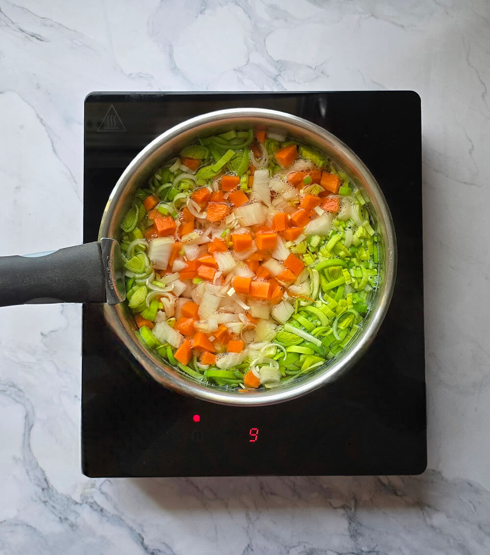 Des dés de carottes et de poireaux émincés cuisant dans une casserole d'eau bouillante sur une plaque à induction.