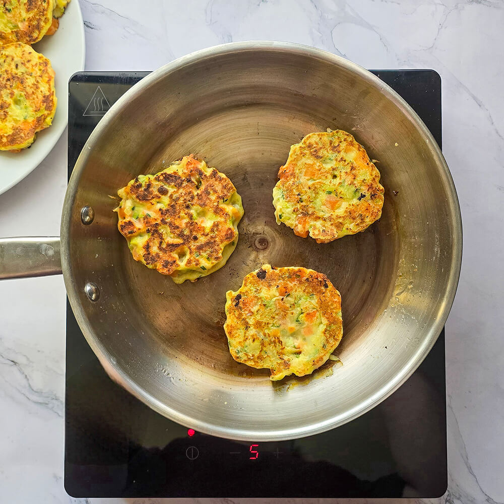 Trois palets de légumes bien dorés en train de dorer dans une poêle en inox sur une plaque de cuisson.