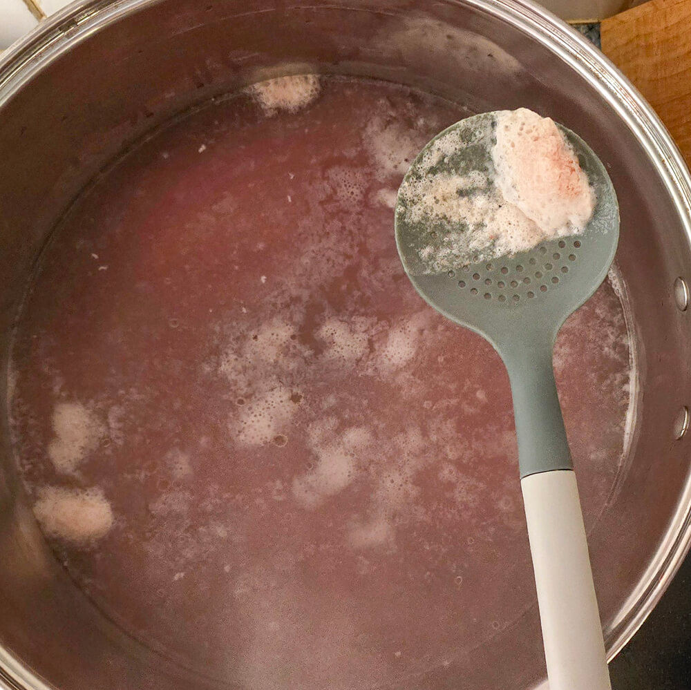 Utilisation d'une écumoire grise pour retirer l'écume et les impuretés à la surface du bouillon de pot-au-feu en cours de cuisson.