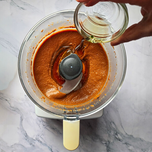 Une main versant de l'huile neutre dans le bol d'un robot pour réaliser une pâté à tartiner.