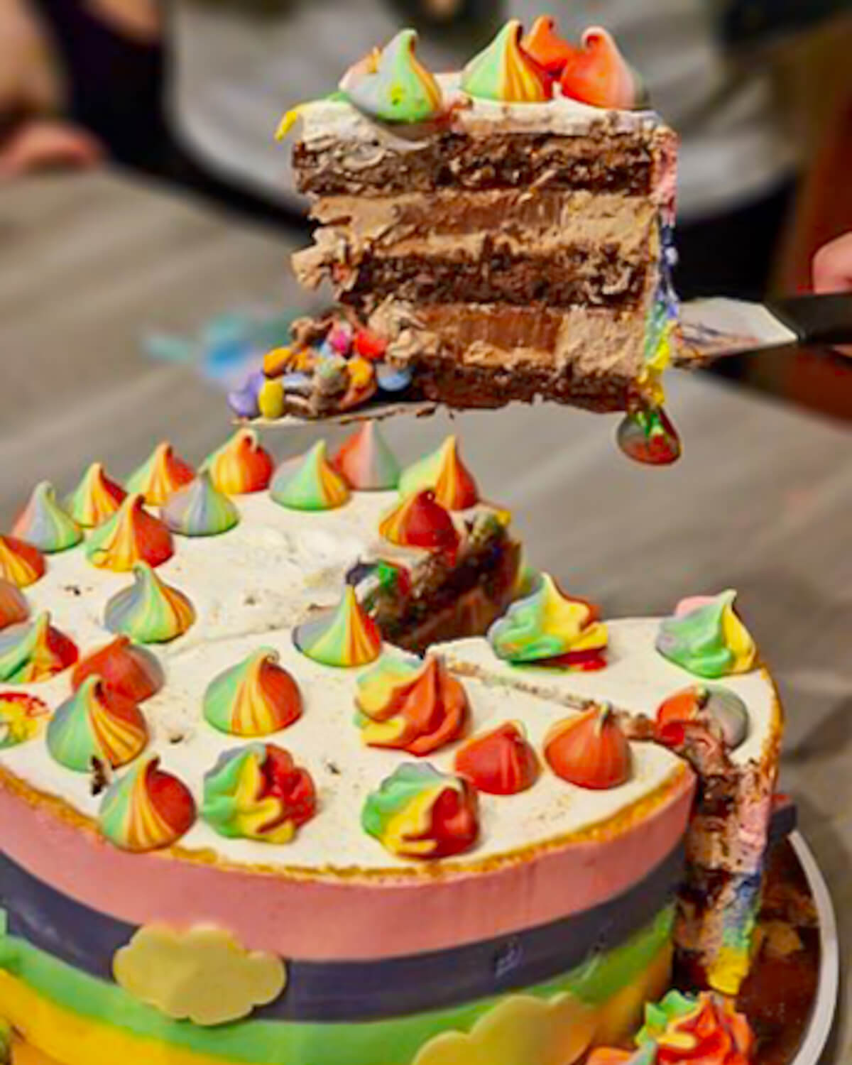 Coupe d'un gâteau d'anniversaire multicouche au chocolat et ganache praliné, révélant une cascade de Smarties colorés à l'intérieur, décoré de meringues arc-en-ciel.