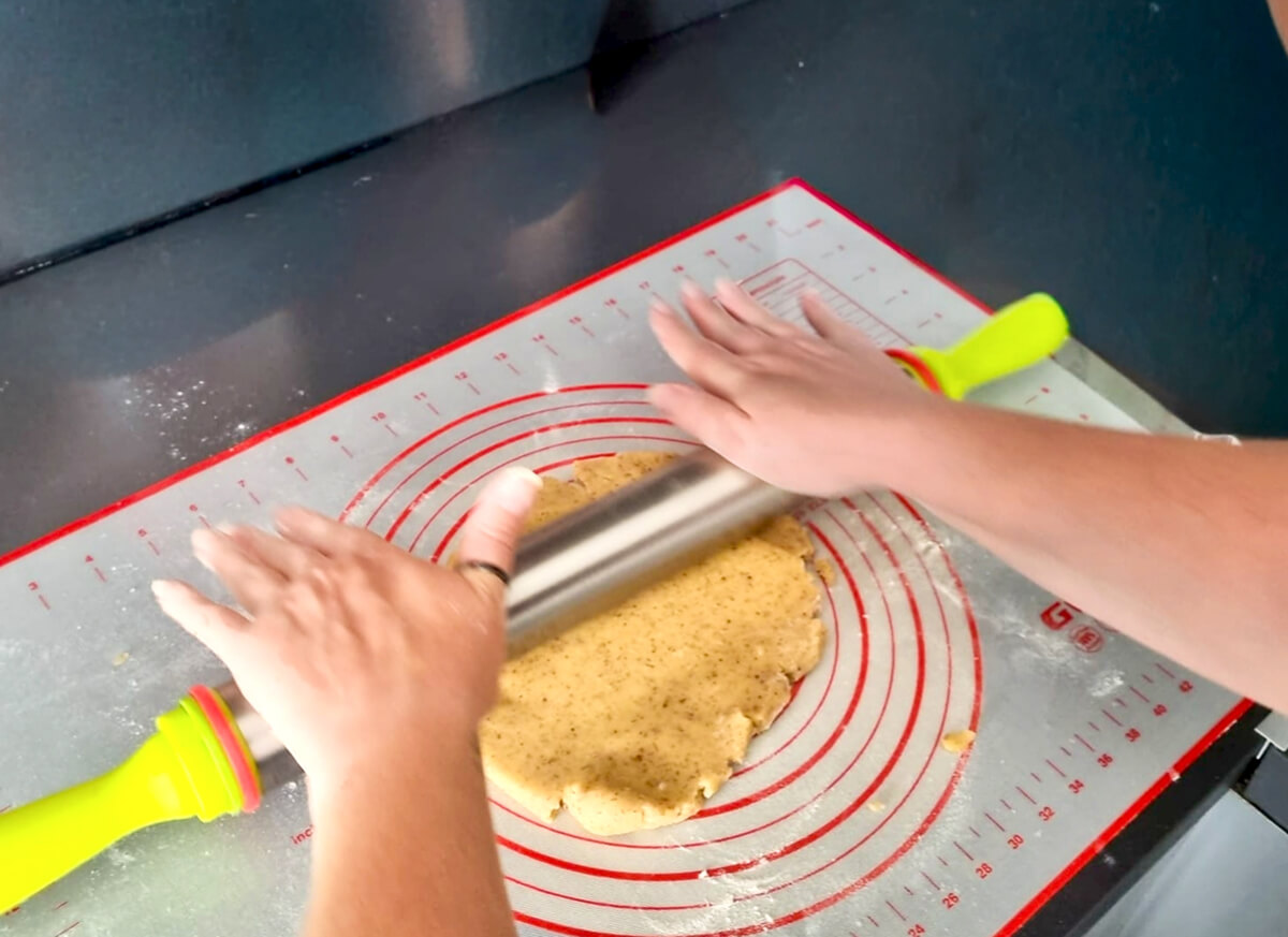 Vue de dessus montrant des mains étalant une pâte à tarte maison avec un rouleau en inox sur un tapis de cuisson gradué.