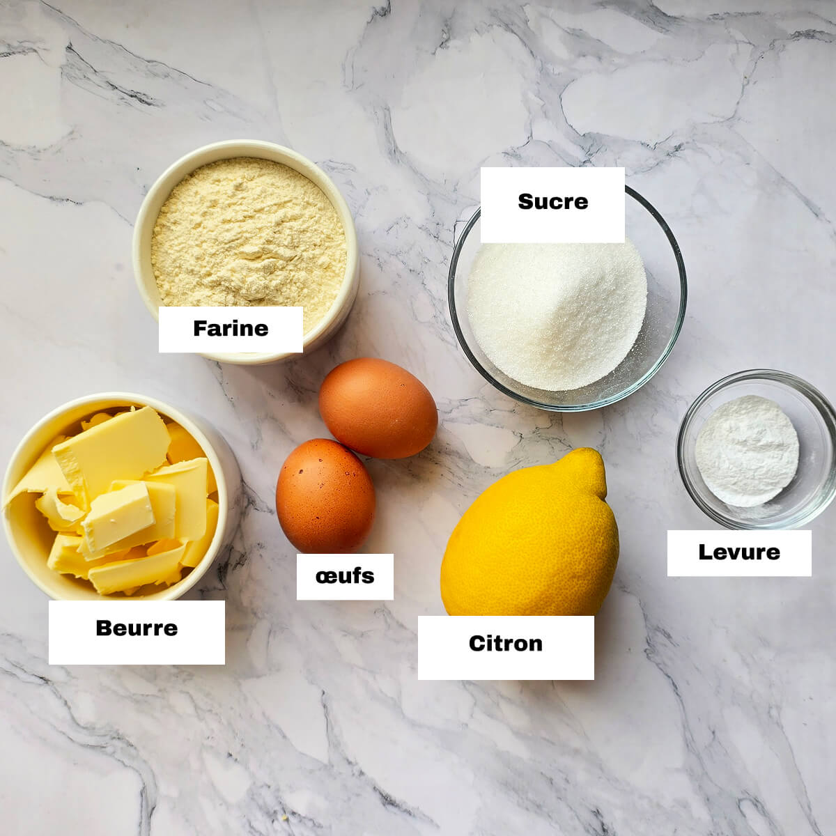 Des ingrédients pour réaliser la recettes des madeleines moelleuses de Cyril Lignac, sur un fond blanc marbré.