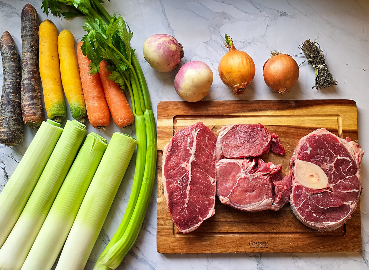 Des ingrédients tels que des légumes, des épices et de la viande pour faire un pot-au-feu traditionnel, sur un fond marbré blanc.