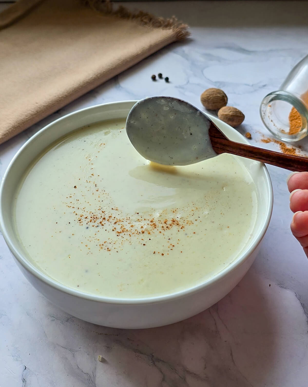 Un bol contenant de la sauce béchamel fait maison, sur fond marbré blanc, noix de muscade, avec une cuillère en bois.