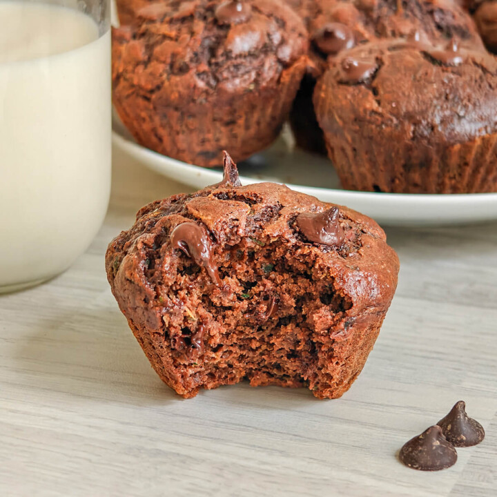 Moitié de muffin chocolat-courgette, verre de lait, et muffins au chocolat en arrière-plan.
