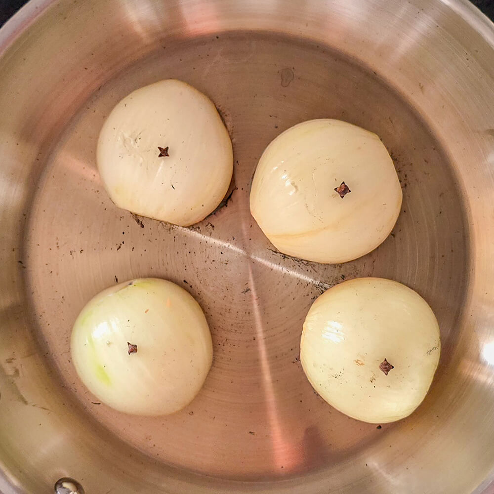 Quatre moitiés d'oignons blancs piqués chacun d'un clou de girofle, placés face coupée contre le fond d'une poêle en inox pour être colorés.