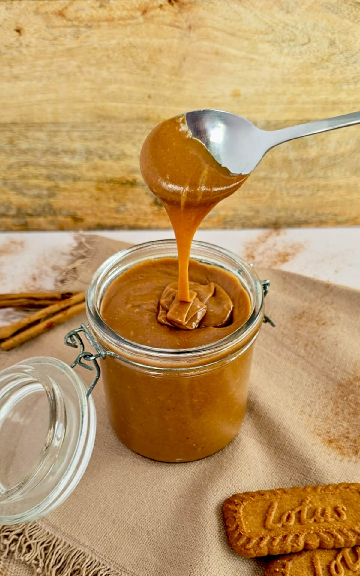 Cuillère soulevant une pâte à tartiner aux spéculoos maison ultra onctueuse au-dessus d'un bocal en verre, avec des biscuits Lotus et de la cannelle en arrière-plan.