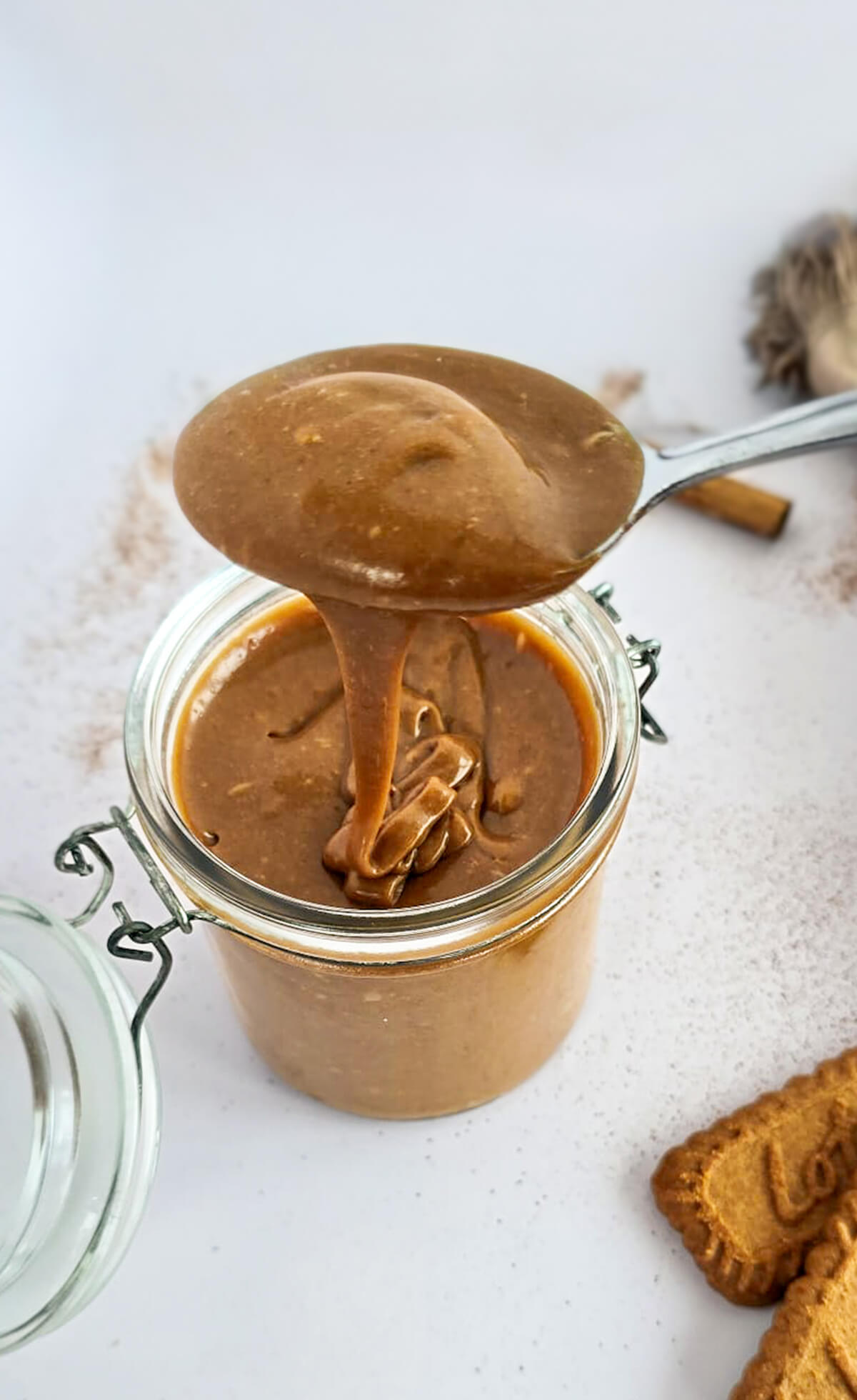 Cuillère soulevant une pâte à tartiner aux spéculoos vegan au-dessus d'un bocal en verre, avec des biscuits Lotus et de la cannelle en arrière-plan.