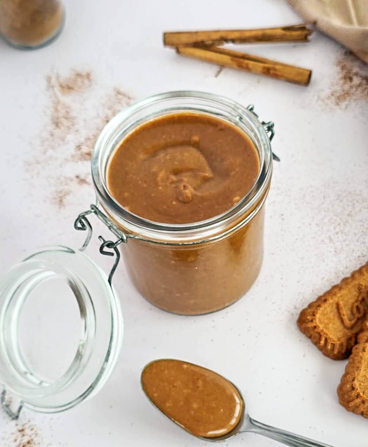 Bocal en verre ouvert de pâte à tartiner aux spéculoos vegan onctueuse avec une cuillère remplie au premier plan, biscuits Lotus et cannelle sur fond blanc.