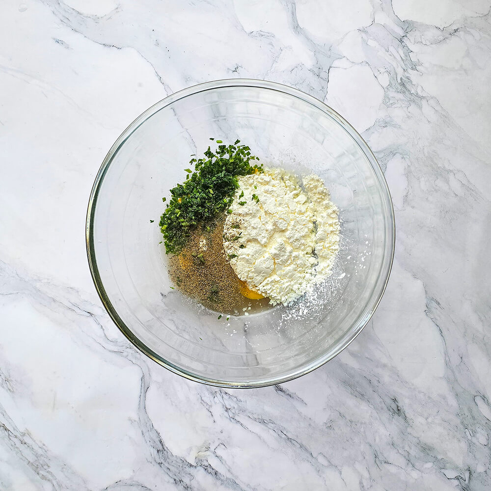 Un saladier en verre contenant de la farine, des œufs, des herbes fraîches hachées et du poivre pour la base de la pâte.