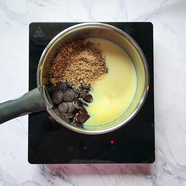 Une casserole sur une plaque de cuisson contenant des pistoles de chocolat noir, de la poudre de noisettes et du lait concentré sucré en cours de préparation.