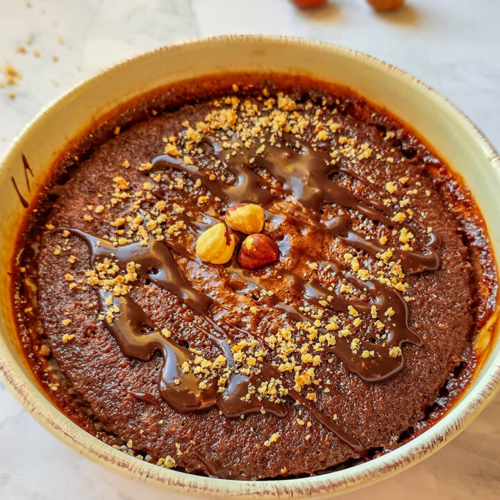 Alt text : Bowl cake au chocolat et noisettes parsemé d'éclats de noisettes et d'un coulis de chocolat fondant, servi dans un bol beige sur un plan de travail en marbre.