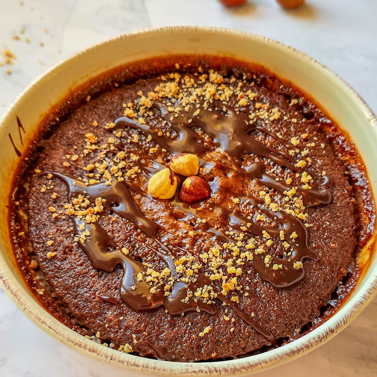 Alt text : Bowl cake au chocolat et noisettes parsemé d'éclats de noisettes et d'un coulis de chocolat fondant, servi dans un bol beige sur un plan de travail en marbre.