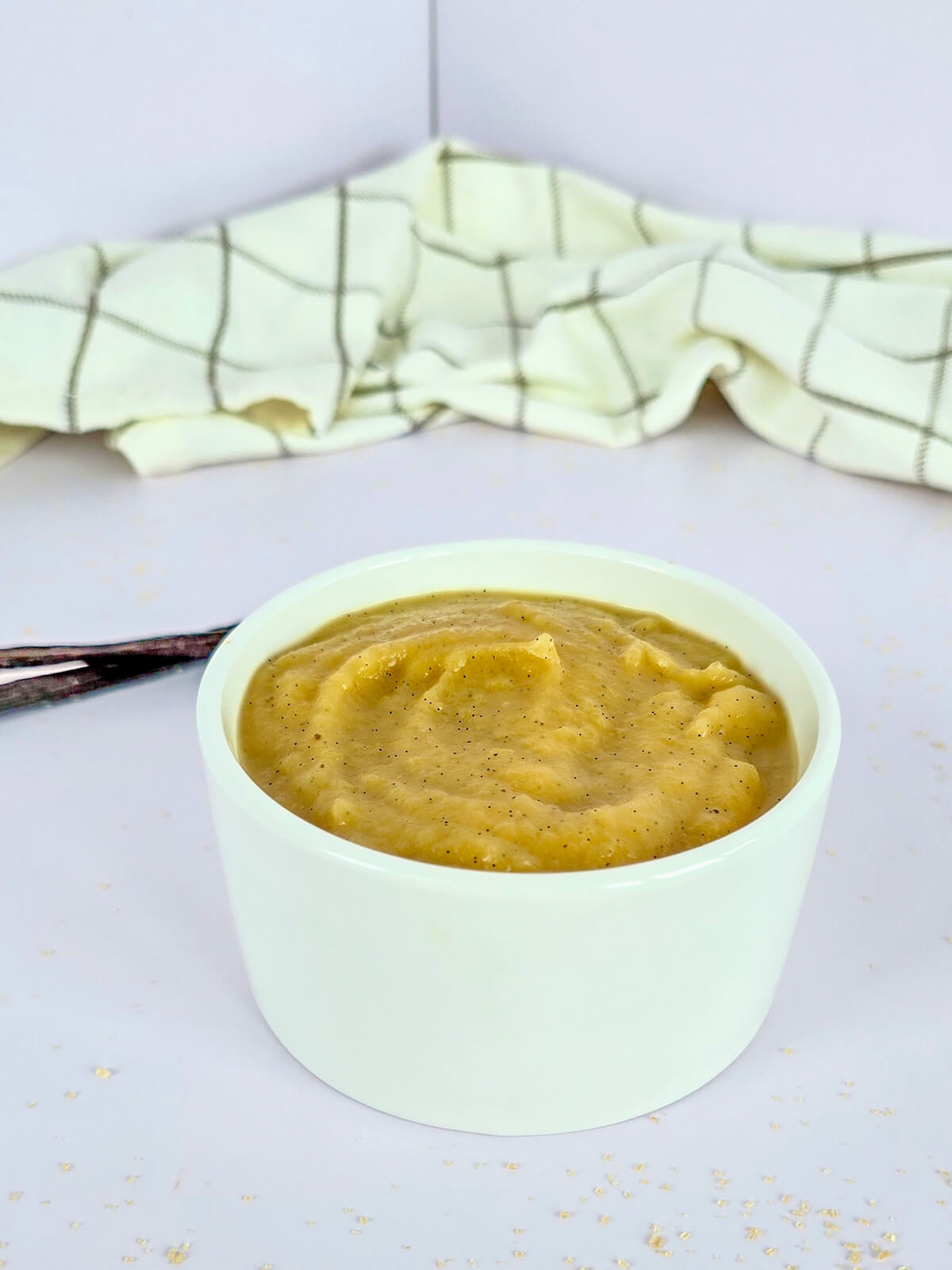Une compote de pommes vanille dans un ramequin blanc, torchon et gousse vanille sur le côté.