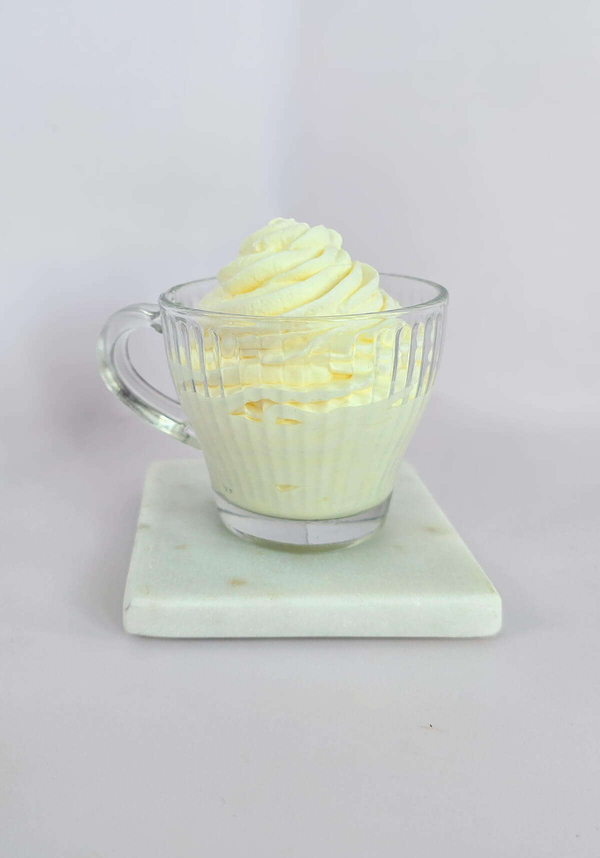 Une tasse en verre remplie de crème chantilly maison ferme et onctueuse, pochée en dôme avec des rainures bien nettes, posée sur un socle en marbre blanc.