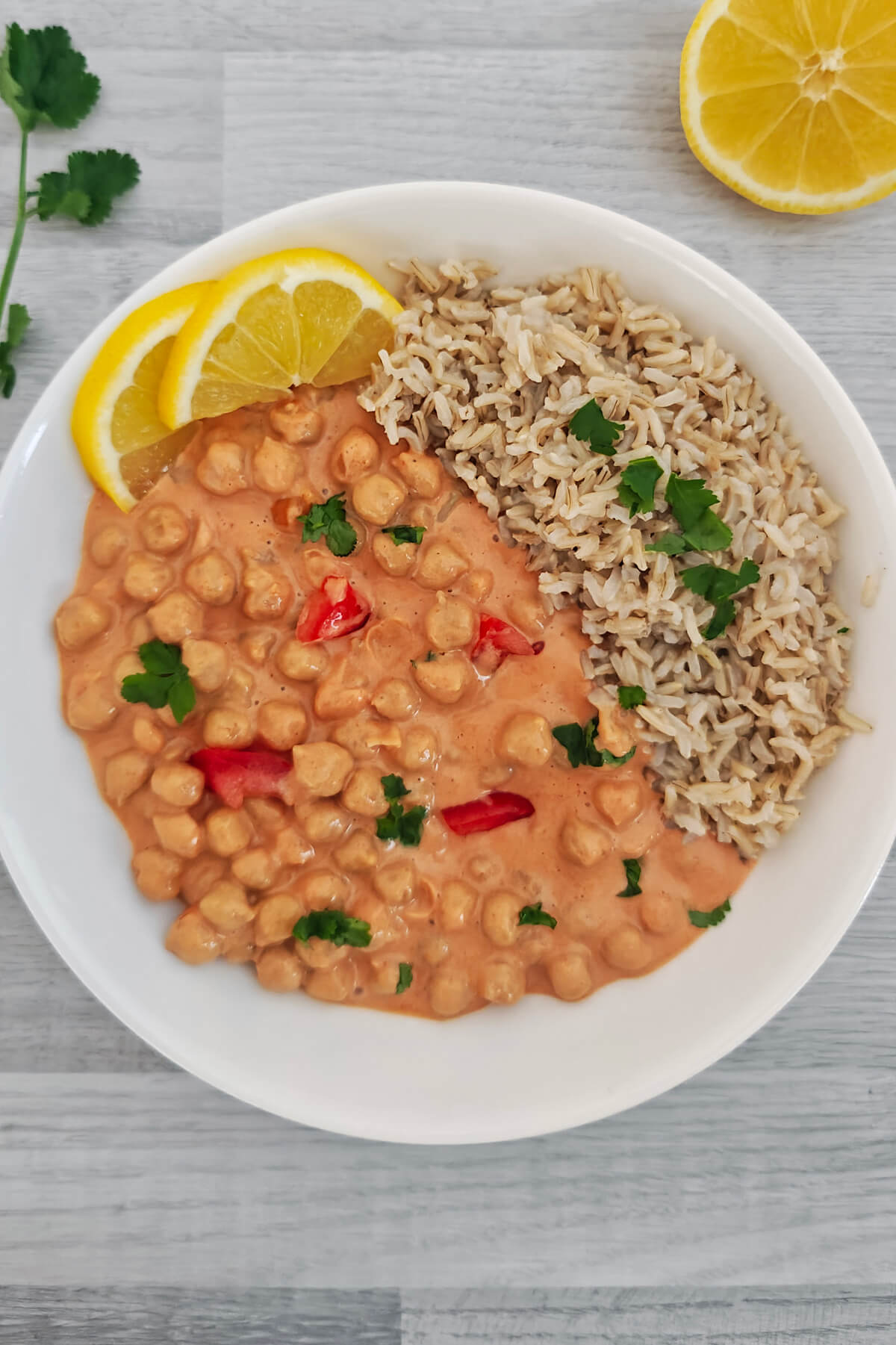 Un bol blanc contenant un curry de pois chiches onctueux au lait de coco et à la tomate, servi avec du riz complet, décoré de tranches de citron jaune et de coriandre fraîche.