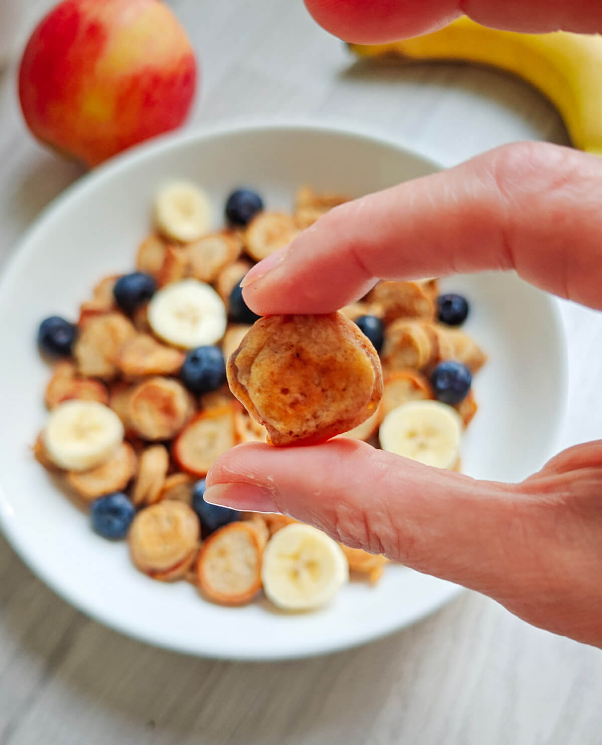 Une main tenant un mini pancake à la banane, assiette de pancakes en arrière-plan.