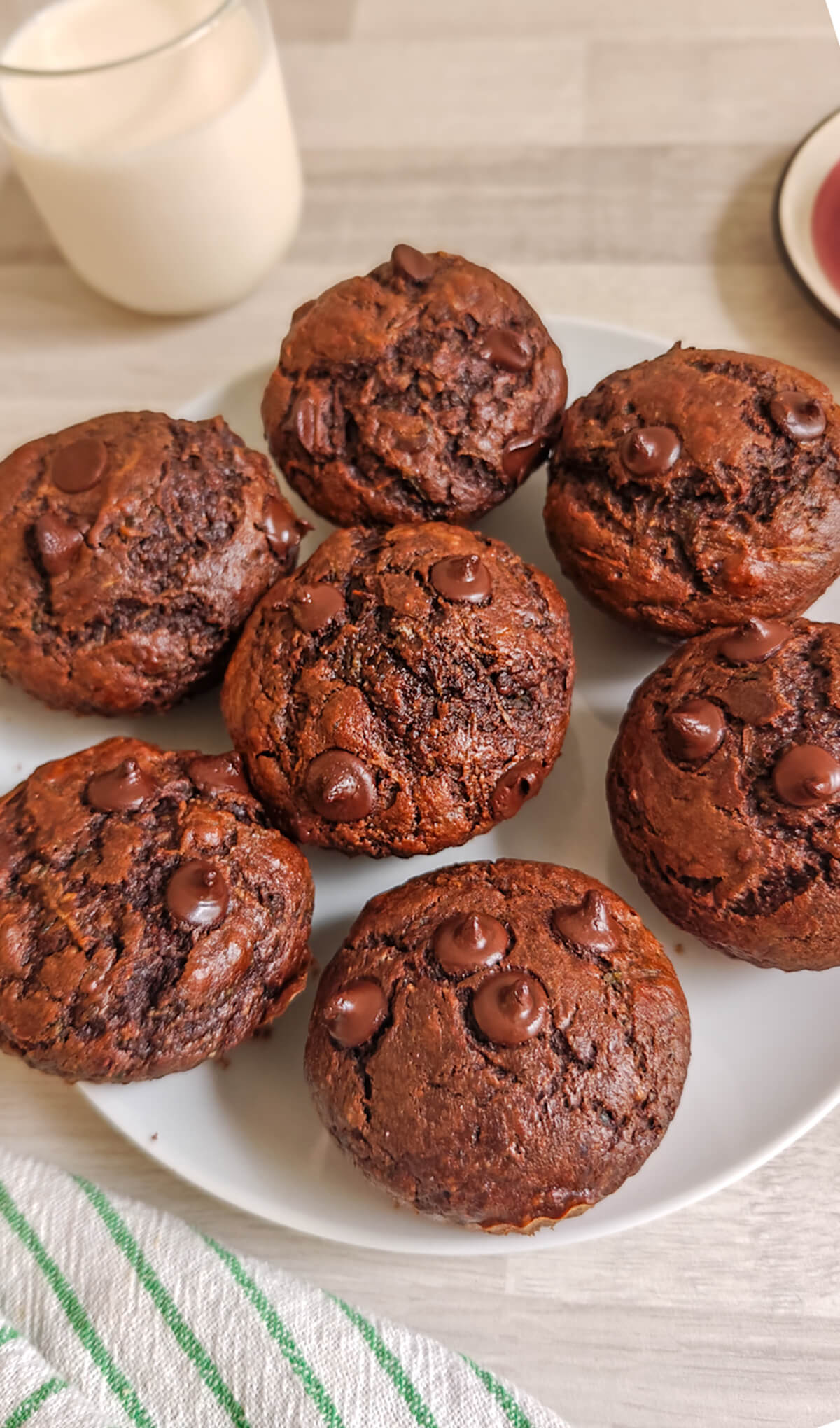 Des muffins tout chocolat courgettes dans une assiette blanche, torchon et verre de lait sur le côté.