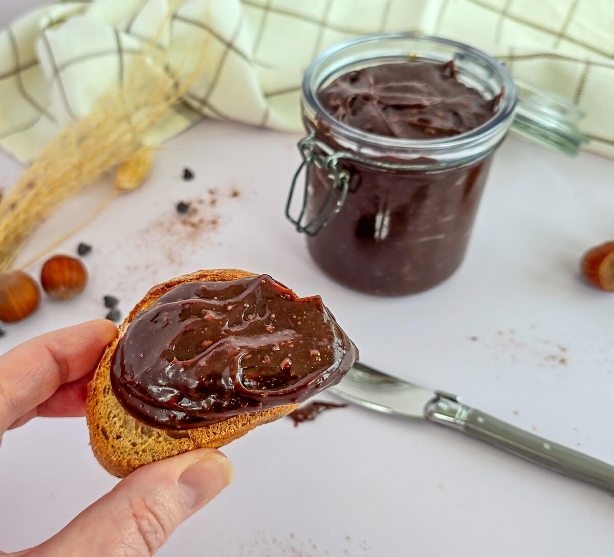 Une main tenant une tartine de pain croustillant généreusement recouverte de pâte à tartiner onctueuse au chocolat noir et éclats de noisettes, avec le bocal en arrière-plan.