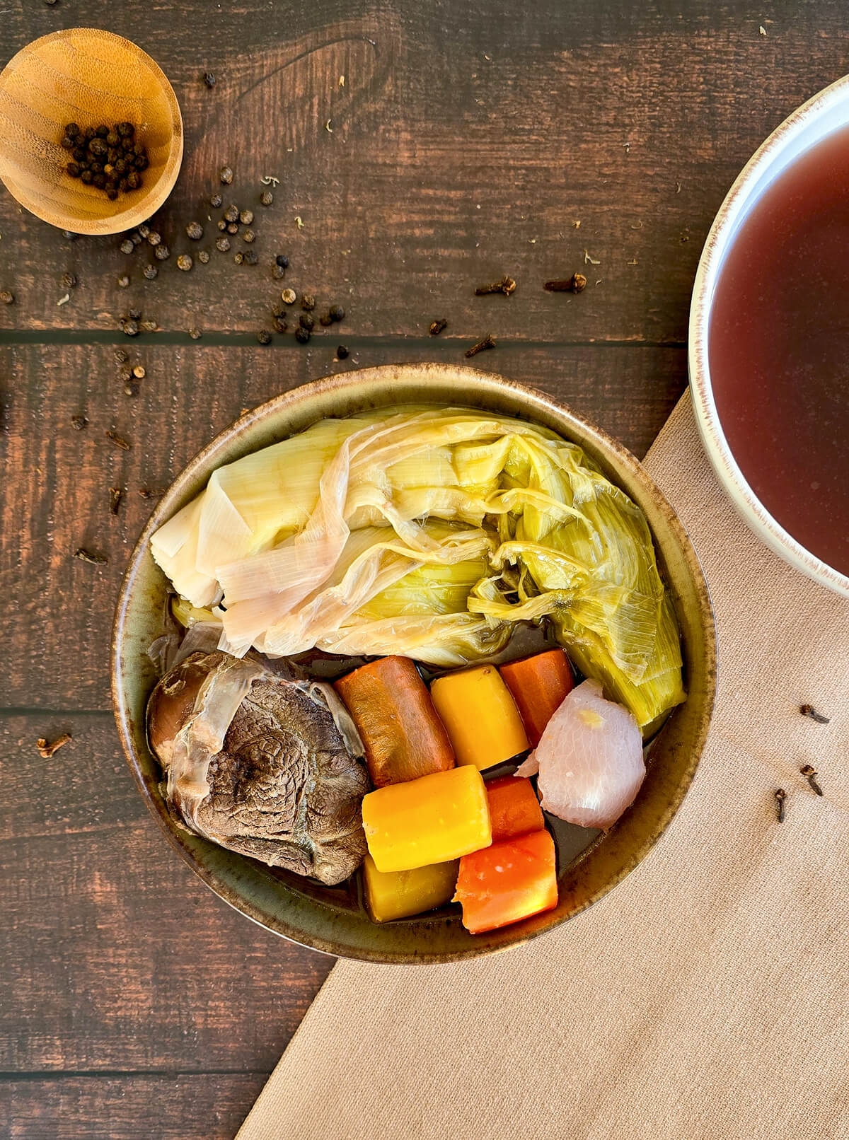 Vue de dessus d'un bol de pot-au-feu traditionnel avec une tranche de bœuf fondante, des poireaux braisés, des carottes jaunes et oranges, servi sur une table en bois avec des grains de poivre et un bol de bouillon.