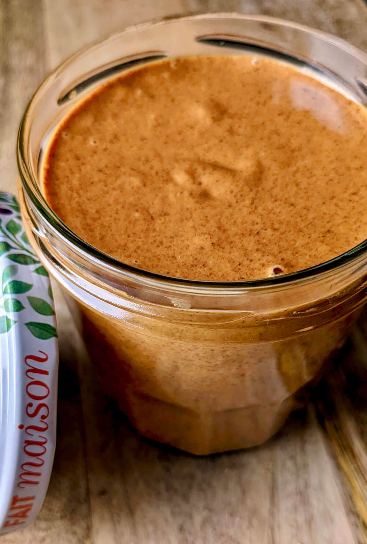 Pot en verre rempli de praliné amande maison onctueux avec un couvercle décoratif 'Fait Maison' posé à côté.