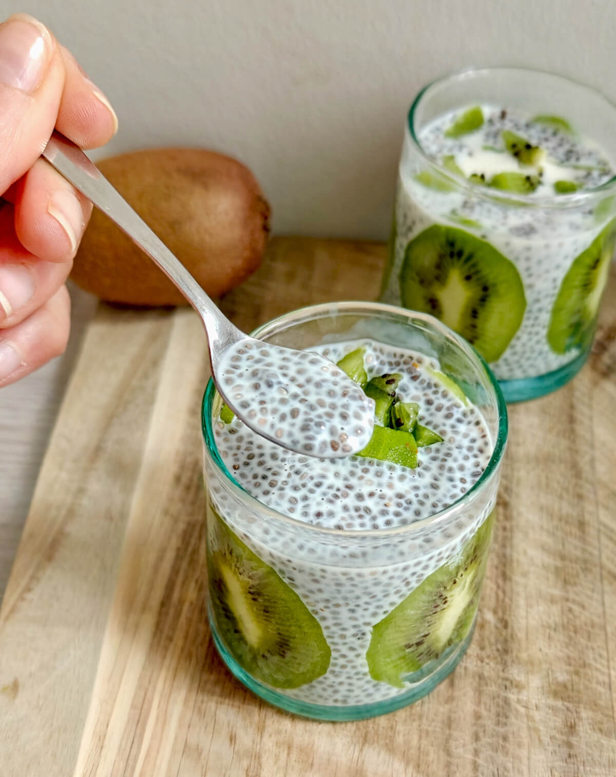 Main tenant une cuillère remplie de chia pudding onctueux au-dessus d'une verrine transparente ornée de tranches de kiwi, illustrant la texture crémeuse de ce petit-déjeuner sain.