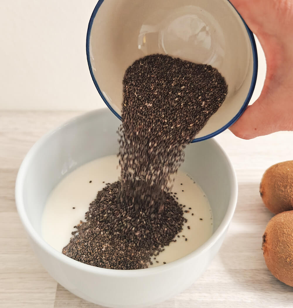 Graines de chia versées d'un bol dans un récipient de lait d'amande pour préparer un pudding de chia maison.