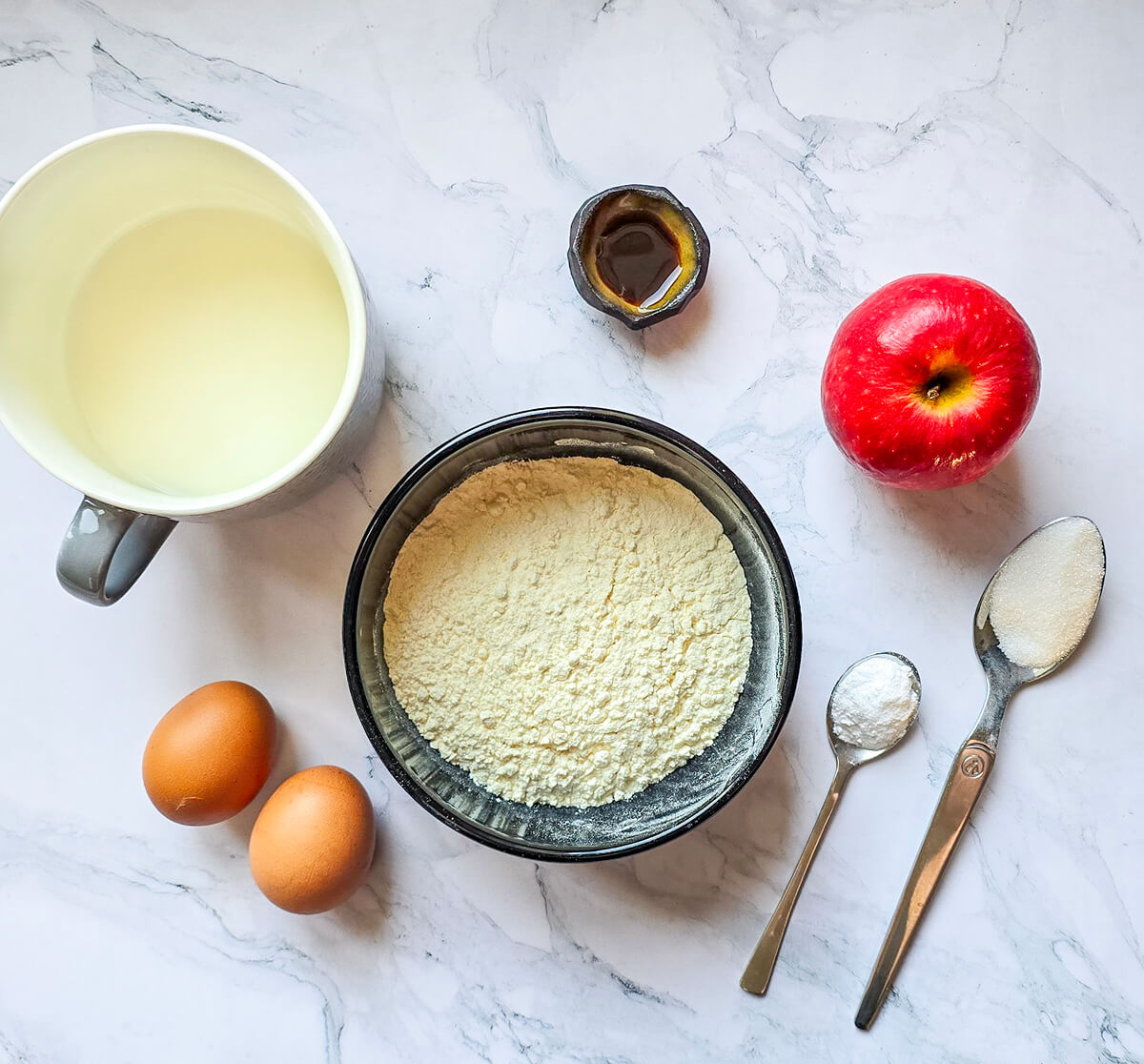 Des ingrédients pour réaliser des pancakes aux pommes fluffy, sur un fond blanc marbré.