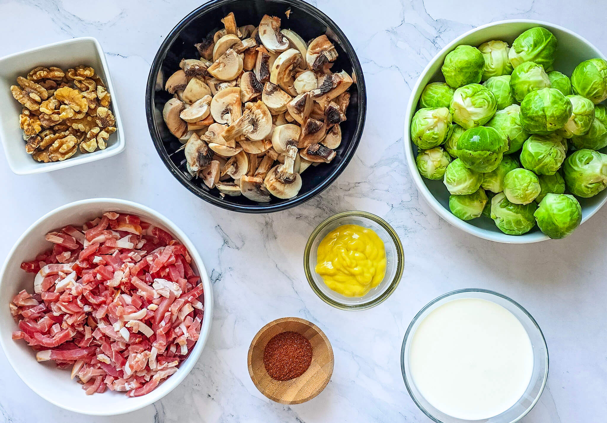 Des ingrédients pour faire une poêlée de choux de Bruxelles, lardons et champignons, sur une surface blanche marbré.