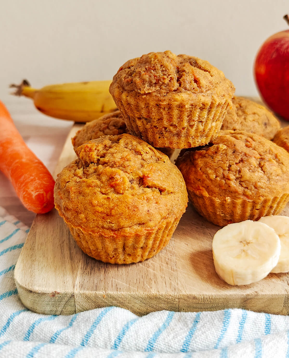 Un plan moyen et appétissant montrant une pile de muffins moelleux aux carottes, pommes et bananes sur une planche à découper en bois. Le muffin du haut montre une texture dorée et rustique, tandis que ceux du bas sont plus lisses. Des tranches de banane fraîche sont posées au premier plan, et l'arrière-plan flou présente les ingrédients entiers : une carotte, une banane entière et une pomme rouge.