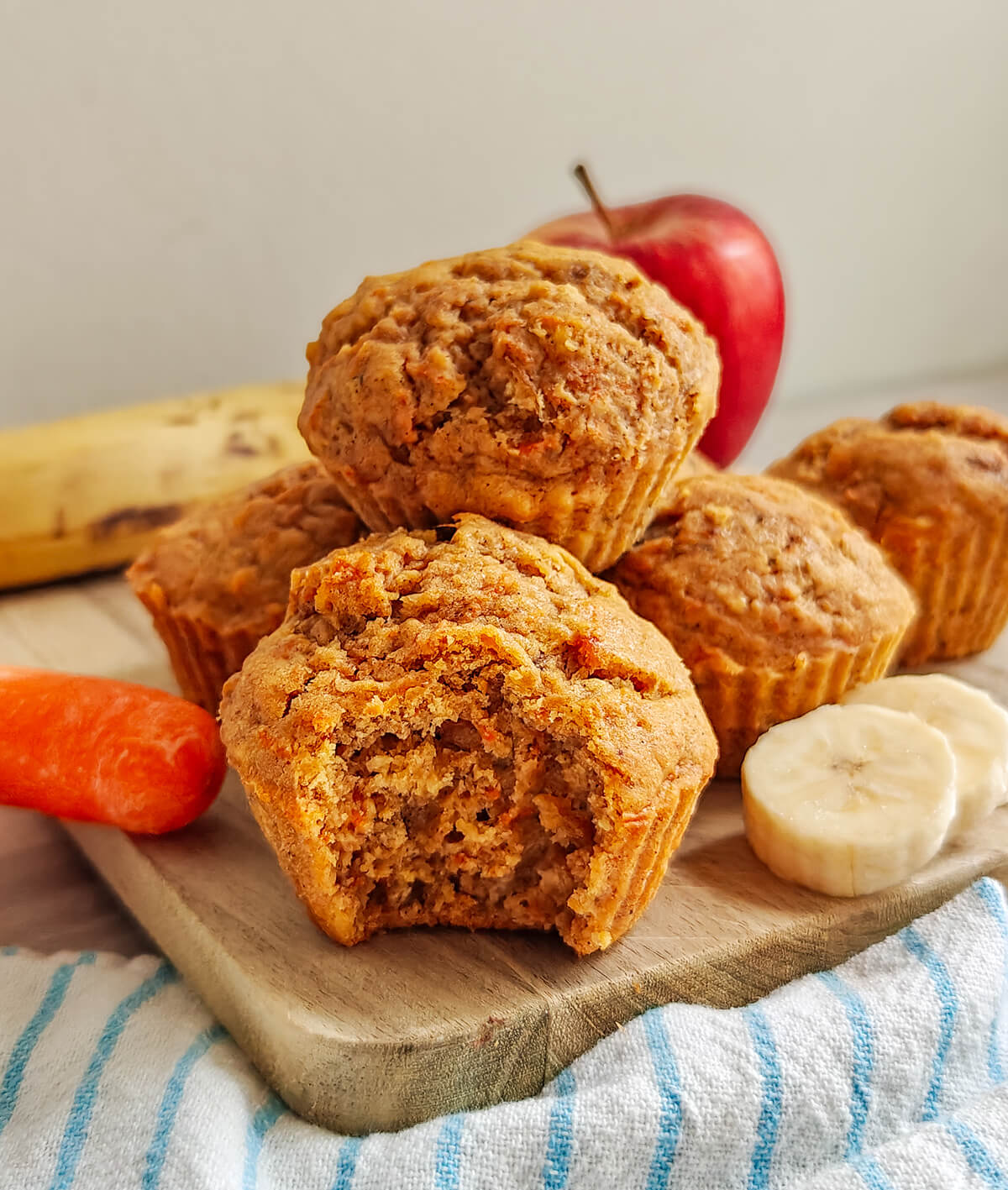 Muffins moelleux aux carottes, pommes et bananes servis sur une planche en bois. Un muffin est croqué pour montrer sa texture aérée à l'intérieur. En arrière-plan, les ingrédients frais sont visibles : une pomme rouge, une banane, des rondelles de banane et une carotte entière