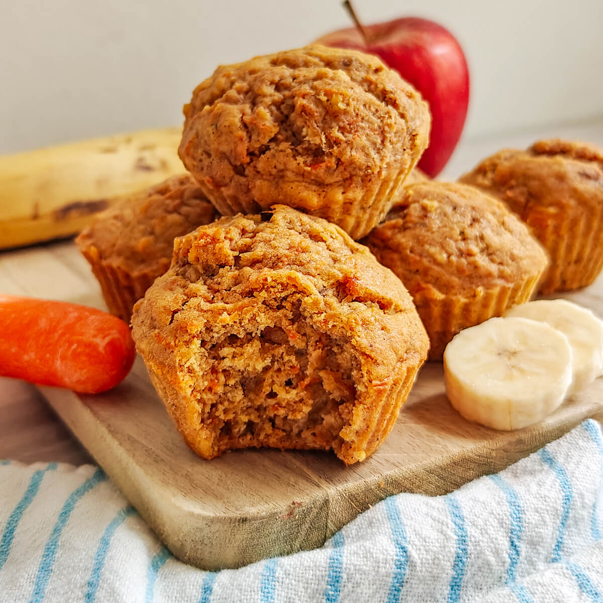 Muffins moelleux aux carottes, pommes et bananes servis sur une planche en bois. Un muffin est croqué pour montrer sa texture aérée à l'intérieur. En arrière-plan, les ingrédients frais sont visibles : une pomme rouge, une banane, des rondelles de banane et une carotte entière
